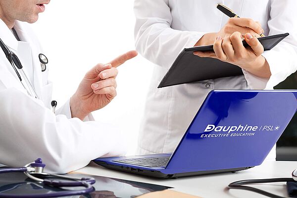 Executive MBA Santé, formation continue de l'Université Paris Dauphine-PSL (visuel principal) Un médecin en blouse avec un stéthoscope travaille avec son assistante à son bureau avec un ordinateur et un bloc notes. Executive MBA Santé, formation continue de l'Université Paris Dauphine-PSL