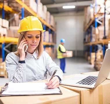 Une professionnelle de la supply chain travaille avec un smartphone et un ordinateur dans un entrepôt rempli de cartons. Programmes de formation continue en Supply Chain et e-commerce de Dauphine Executive Education