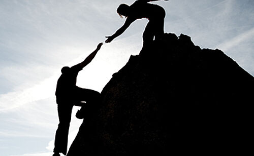 Une femme aide un homme à gravir une montagne. Photo en contre jour symbolisant l'accompagnement et le coaching