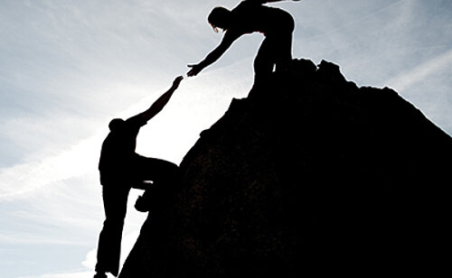 Une femme aide un homme à gravir une montagne. Photo en contre jour symbolisant l'accompagnement et le coaching