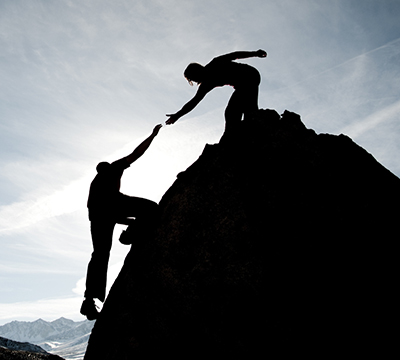 Une femme aide un homme à gravir une montagne. Photo en contre jour symbolisant l'accompagnement et le coaching